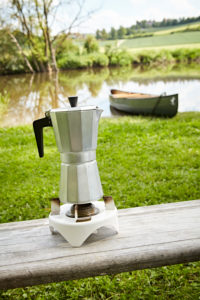 Espressokanne auf einem Campingkocher, im Hintergrund ein Kanu im Fluss Espressokanne auf einem Campingkocher, im Hintergrund ein Kanu im Fluss