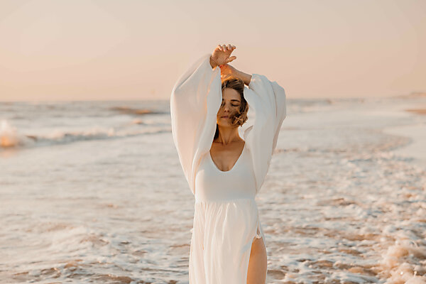 Bildagentur | mauritius images | Woman in a flowing white dress standing in ocean waves at sunset Bildagentur | mauritius images | Woman in a flowing white dress standing in ocean waves at sunset