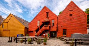 Tourists walk in front of a sculptural wooden house with wooden staircase, surrounded by a paved courtyard on the tables and benches, decorated with flower tubs, in Bryggen, Bergen, Hordaland, Norway, Scandinavia, Europe Tourists walk in front of a sculptural wooden house with wooden staircase, surrounded by a paved courtyard on the tables and benches, decorated with flower tubs, in Bryggen, Bergen, Hordaland, Norway, Scandinavia, Europe