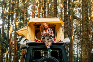 Travel and wanderlust lifestyle concept with happy lonely adult woman sit down on the roof tent car vehicle with wood forest in background enjoying nature and outdoors vacation Travel and wanderlust lifestyle concept with happy lonely adult woman sit down on the roof tent car vehicle with wood forest in background enjoying nature and outdoors vacation