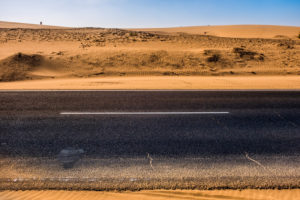 Black asphalt road with sand dunes and desert on left and right and blue sky in background - travel concept in arid desertic world due a bad climate change future - water emergency planet Black asphalt road with sand dunes and desert on left and right and blue sky in background - travel concept in arid desertic world due a bad climate change future - water emergency planet