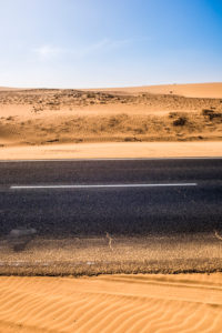 Black asphalt road with sand dunes and desert on left and right and blue sky in background - travel concept in arid desertic world due a bad climate change future - water emergency planet Black asphalt road with sand dunes and desert on left and right and blue sky in background - travel concept in arid desertic world due a bad climate change future - water emergency planet