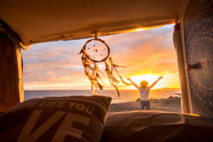 Travel and freedom concept with standing happy joy woman outside vehicle van enjoy the sunset at the beach - dreamcatcher in foreground and sun in background Travel and freedom concept with standing happy joy woman outside vehicle van enjoy the sunset at the beach - dreamcatcher in foreground and sun in background