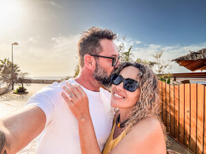 Mature couple enjoying a summer vacation at a tropical beach, taking a selfie with a smartphone under a sunny sky, surrounded by white sand, clear blue ocean waves, and warm sunlight