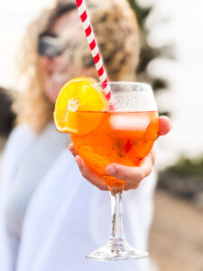 An elegant summer scene featuring a smiling woman offering an orange spritz cocktail with ice cubes and a straw, set in a bright, vibrant atmosphere perfect for socializing