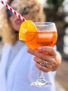 A smiling woman holds an orange spritz cocktail toward the camera, with ice cubes and a straw, set in a vibrant, cheerful, and elegant summer environment perfect for leisure and social moments