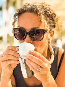 A close-up portrait of a beautiful mature woman sitting outdoors after fitness, relaxing in sunlight, holding an espresso cup, wearing sporty casual clothes, sunglasses, and white headphones around her neck