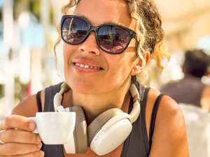 Portrait of a stylish mature woman relaxing outdoors after a workout, wearing sporty casual attire, sunglasses, and white headphones, enjoying an espresso coffee in sunlight, with a confident expression