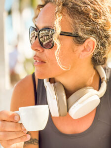 athletic woman relaxing outdoors in sunlight after a fitness session, wearing sporty casual clothes, sunglasses, white headphones, and holding an espresso coffee in her hand with a confident expression