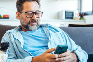 Pensive serious young man sit on sofa at home text message on modern smartphone gadget. Focused genx male look at cellphone screen browse surf wireless internet on device. Technology concept
