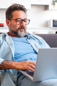 An adult man wearing glasses and a beard sitting at home on a sofa in a relaxed position, using a laptop to read email content with bad news, appearing serious, thoughtful, and focused