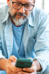 A thoughtful man seated on his sofa during the evening at home, holding a cellphone in one hand, scrolling through notifications or typing a message while appearing serious and focused