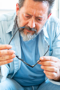 A middle-aged man sitting alone on a couch in his home, thoughtfully checking his eyeglasses, reflecting on eyesight problems and middle-age vision correction needs
