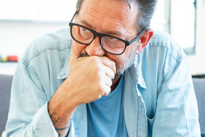A mature man with glasses and a beard sitting alone on a sofa in his living room, touching his chin with a serious and introspective expression, thinking deeply about family and relationship struggles