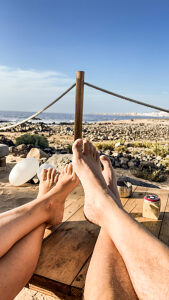 POV of a couple on vacation showing their legs and feet with a scenic tropical beach, ocean, and sky in the background, illustrating a relaxing and romantic getaway to an exotic destination