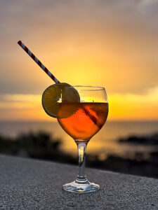A large glass of orange spritz cocktail with a slice of orange, set against a warm tropical sunset, highlighting the vibrant colors of the sky and drink. Summer vacation concept lifestyle