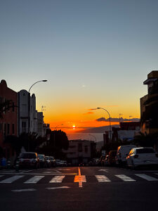 A scenic road with parked cars, ocean view, and sunset colors on the horizon, featuring a pedestrian crosswalk in the foreground and vibrant shadows. Sunlight amazing sky in background