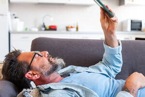 Man lying on the sofa at home in a relaxed position, using his phone to send messages to friends, scrolling through entertaining media content and videos while enjoying his leisure time