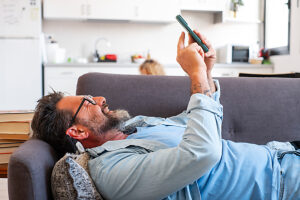 Man lying on the sofa at home in a relaxed position, using his phone to send messages to friends, scrolling through entertaining media content and videos while enjoying his leisure time