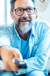 A happy adult man sitting cheerful on a sofa in front of the television, holding a remote control, watching and changing channels, smiling and enjoying their leisure time at home