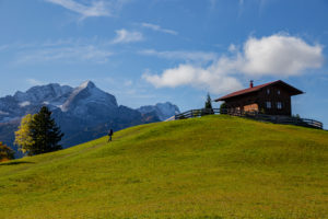Man walks on meadow towards mountain hut at Eckbauer in autumn, in the background the Wetterstein Mountains with Alpspitze and Zugspitze, blue sky, clouds, Garmisch-Partenkirchen, Upper Bavaria, Bavaria, southern Germany, Germany Man walks on meadow towards mountain hut at Eckbauer in autumn, in the background the Wetterstein Mountains with Alpspitze and Zugspitze, blue sky, clouds, Garmisch-Partenkirchen, Upper Bavaria, Bavaria, southern Germany, Germany
