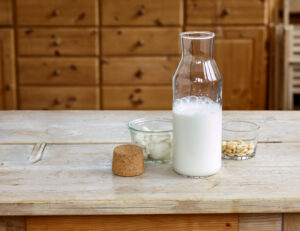 Steps for making almond milk, finished almond milk in a glass bottle with cork and a glass bowl with blanched almonds and a glass bowl with the squeezed almond paste, on a wooden table