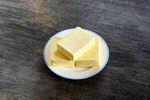 ayurvedic diet, steps to prepare homemade ghee from butter - three pieces of organic sour cream butter on a white porcelain plate, on a dark wooden tabletop