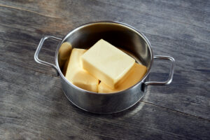 ayurvedic diet, steps to prepare homemade ghee from butter - three pieces of organic sour cream butter in a stainless steel cooking pot on a dark wooden tabletop