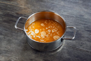 ayurvedic diet, steps to prepare homemade ghee from butter - melted sour cream butter in a stainless steel saucepan, on a dark wooden tabletop, on the surface begins to settle egg white.