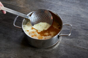 ayurvedic diet, steps to prepare homemade ghee from butter - melted sour cream butter in a stainless steel saucepan on a dark wooden tabletop, the egg white foam is skimmed off with a ladle