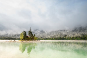 A small lonely island with a log cabin and red roof, in the middle of the beautiful Eibsee below the Zugspitze in the Bavarian Alps of the Wetterstein Mountains during the Eisheiligen - fresh snow in the background on the forest and cloud mood. A small lonely island with a log cabin and red roof, in the middle of the beautiful Eibsee below the Zugspitze in the Bavarian Alps of the Wetterstein Mountains during the Eisheiligen - fresh snow in the background on the forest and cloud mood.