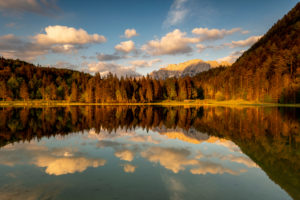 Evening light with light clouds at the Ferchensee, above Mittenwald. The edge of the forest and the Karwendel in the background are warmly lit and reflected in the clear water of the lake. Evening light with light clouds at the Ferchensee, above Mittenwald. The edge of the forest and the Karwendel in the background are warmly lit and reflected in the clear water of the lake.