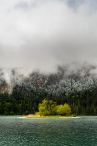 Small green island in the Eibsee, below the Zugspitze in the Bavarian Alps of the Wetterstein Mountains during the ice saints in spring. In the background snow on the fresh green of the forest and thick rain clouds. Small green island in the Eibsee, below the Zugspitze in the Bavarian Alps of the Wetterstein Mountains during the ice saints in spring. In the background snow on the fresh green of the forest and thick rain clouds.
