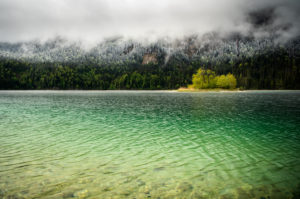 Small green island in the Eibsee, below the Zugspitze in the Bavarian Alps of the Wetterstein Mountains during the ice saints in spring. In the background snow on the fresh green of the forest and thick rain clouds. Small green island in the Eibsee, below the Zugspitze in the Bavarian Alps of the Wetterstein Mountains during the ice saints in spring. In the background snow on the fresh green of the forest and thick rain clouds.