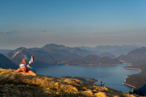 Trailrunnerin Katharina Kirschner liegt auf einer Frühlingswiese am Simetsberg, im Hintergrund der Walchensee und lächelt in die Kamera. Trailrunnerin Katharina Kirschner liegt auf einer Frühlingswiese am Simetsberg, im Hintergrund der Walchensee und lächelt in die Kamera.