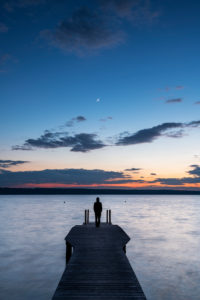 Ein einzelner Mann mit Kapuzenpullover steht auf einem Holzsteg am Ufer des Ammersee während Sonnenuntergang mit Sichelmond. Ein einzelner Mann mit Kapuzenpullover steht auf einem Holzsteg am Ufer des Ammersee während Sonnenuntergang mit Sichelmond.