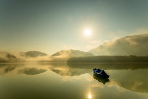A single rowing boat with tarpaulin is moored at the Sylvensteinspeicher lake, while the sun sends its rays over the lake with light fog in the early morning and is reflected in the water. A single rowing boat with tarpaulin is moored at the Sylvensteinspeicher lake, while the sun sends its rays over the lake with light fog in the early morning and is reflected in the water.