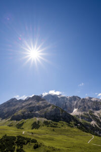 The famous Karwendelhaus in the Karwendel, in the Tyrolean Alps under blue sky and sun with sun star. In the foreground the pastures of the Graßlegerbichl, middle ground dominated by the local mountain of the hut, the Hochalmkreuz (2192m) and in the background Schlauchkakopf (2500m), Birkkarspitze (2749m) and the Ödkarspitzen.