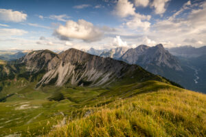 Late summer day with view from Schönalmspitze to Fleischbank, in the foreground golden grass and a mountain meadow, while the ridge leads to the next peak and light clouds shade parts of the picture.