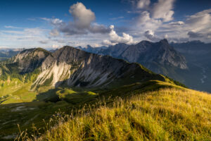Late summer day with view from Schönalmspitze to Fleischbank, in the foreground golden grass and a mountain meadow, while the ridge leads to the next peak and light clouds shade parts of the picture.