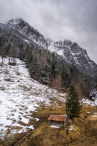 A single haystack with red roof above the Ferchensee near Mittenwald in the Bavarian Alps in spring. A still partly snow-covered mountain meadow, with forest and snow, in the background the massive Wetterstein mountains with the Wetterstein in clouds and overcast sky.