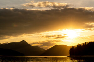 Evening mood shortly before sunset at Walchensee in the Bavarian Alps, in the foreground an angler in a rowing boat, in the background the Simetsberg, cloudy atmosphere and the setting sun.