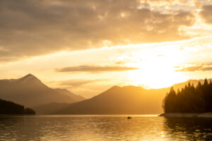 Evening mood shortly before sunset at Walchensee in the Bavarian Alps, in the foreground an angler in a rowing boat, in the background the Simetsberg, cloudy atmosphere and the setting sun.