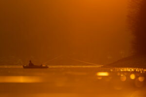 An angler sits in his boat at Walchensee during evening backlight, his fishing rod and fishing line pointing towards the shore, while the water glitters and the whole sznerie is bathed in reddish orange evening light.