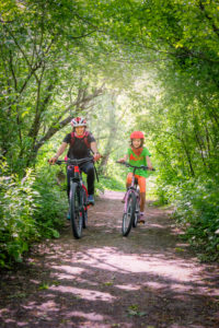 Italy, Veneto, Belluno, Agordino, smiling mom and daughter ride a bicycle down a road among the trees Italy, Veneto, Belluno, Agordino, smiling mom and daughter ride a bicycle down a road among the trees