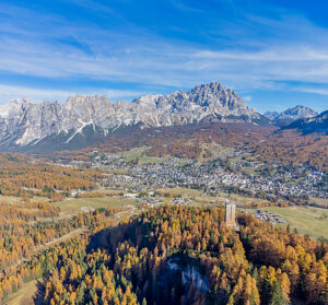 Aerial view of the Sacrario Militare di Pocol surrounded by golden larch trees, with Cortina d'Ampezzo town center in front of the iconic Pomagagnon and Monte Cristallo mountains in the background, during autumn. Cortina d'Ampezzo, Province of Belluno, Veneto, Italy