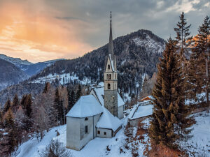 Aerial view of the Church of San Simon in Vallada Agordina, covered in snow at sunset, historic monument in the province of Belluno, Veneto region, northern Italy