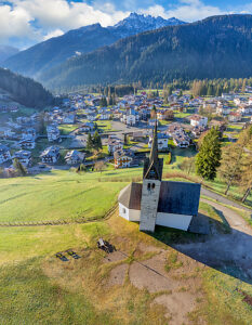 Aerial view of the Church of the Beata Vergine della Salute on the hill overlooking the village of Caviola, with its rooftops and houses nestled among alpine meadows and forest. Valle del Biois, Falcade, Belluno province, Veneto region, northeastern Italy
