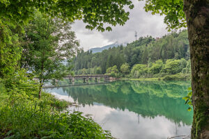 View of the old concrete bridge crossing Lake Centro Cadore, framed by lush lakeside trees. Domegge di Cadore, Belluno province, Veneto, Italy
