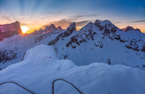 Aerial winter view of Passo Giau at dawn, with the sun rising behind Forcella Giau and Monte Cernera visible on the right. Snow-covered alpine slopes glow in the early morning light. Dolomites, Colle Santa Lucia municipality, Belluno province, Veneto region, Italy